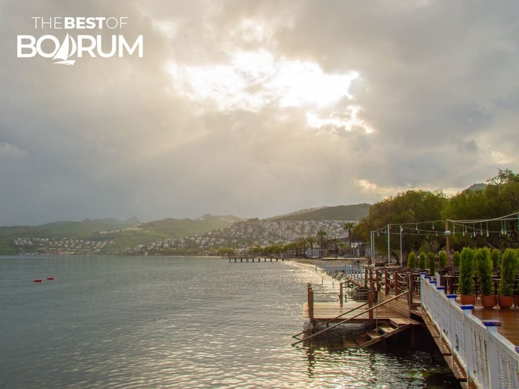 Rainy day on the Ortakent coast of Bodrum, with clouds covering the sky, a few sunbeams breaking through, and a dramatic atmosphere.