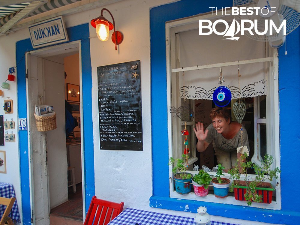 A lady, the restaurant owner, smiling and waving from the window of a Bodrum restaurant with blue painted window and door frames. Tipping in Turkey is based on the hospitality.