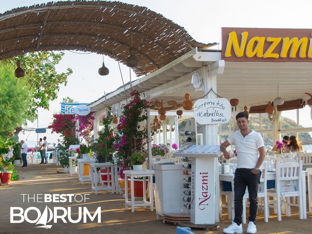 A waiter smiling at the camera in Gumusluk, with colourful flowers and a dirt road in the background.