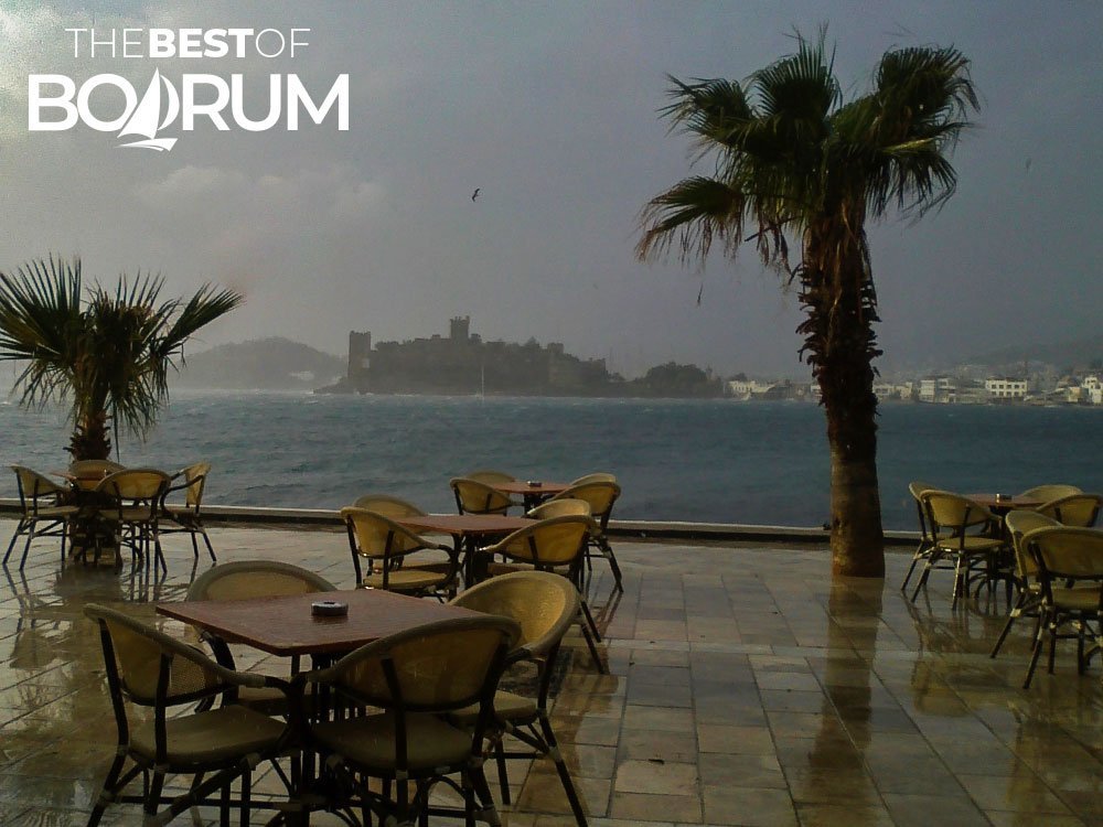Rainy day in winter in Bodrum, empty cafe tables under rain, view of the Aegean Sea and Bodrum Castle.