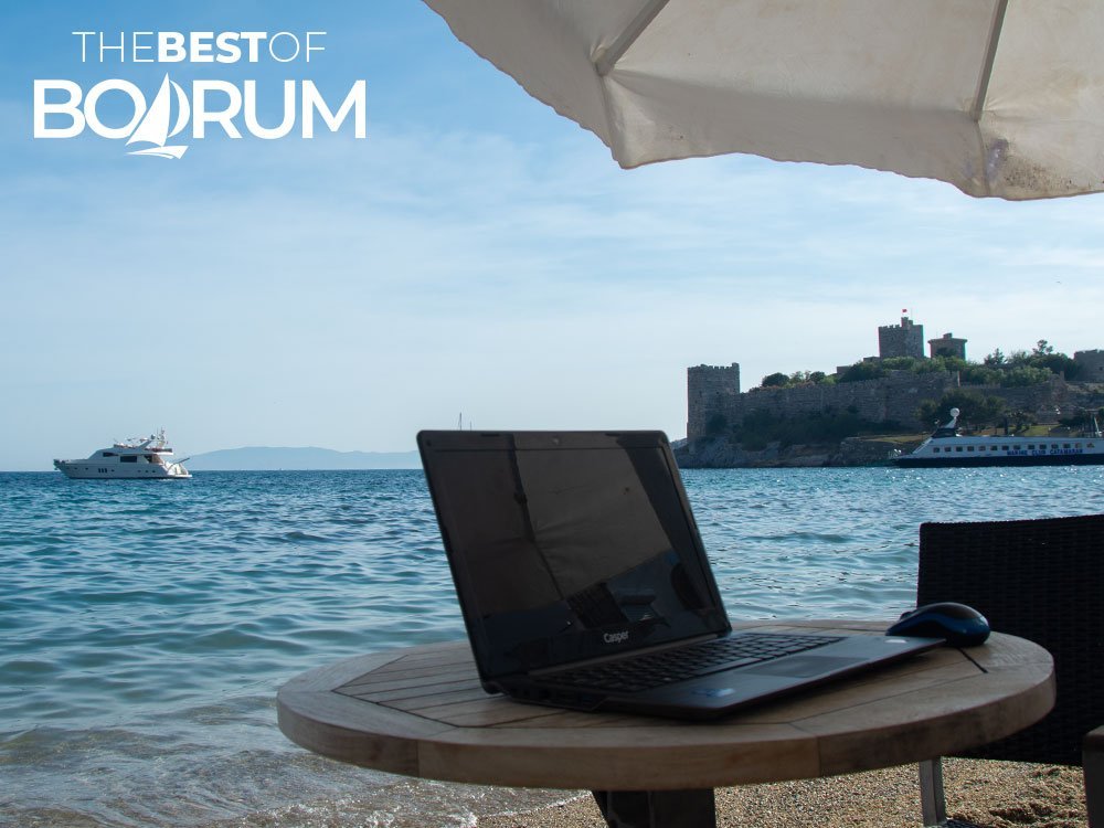 A laptop on a seaside table facing Bodrum Castle, representing travellers sharing their own stories.